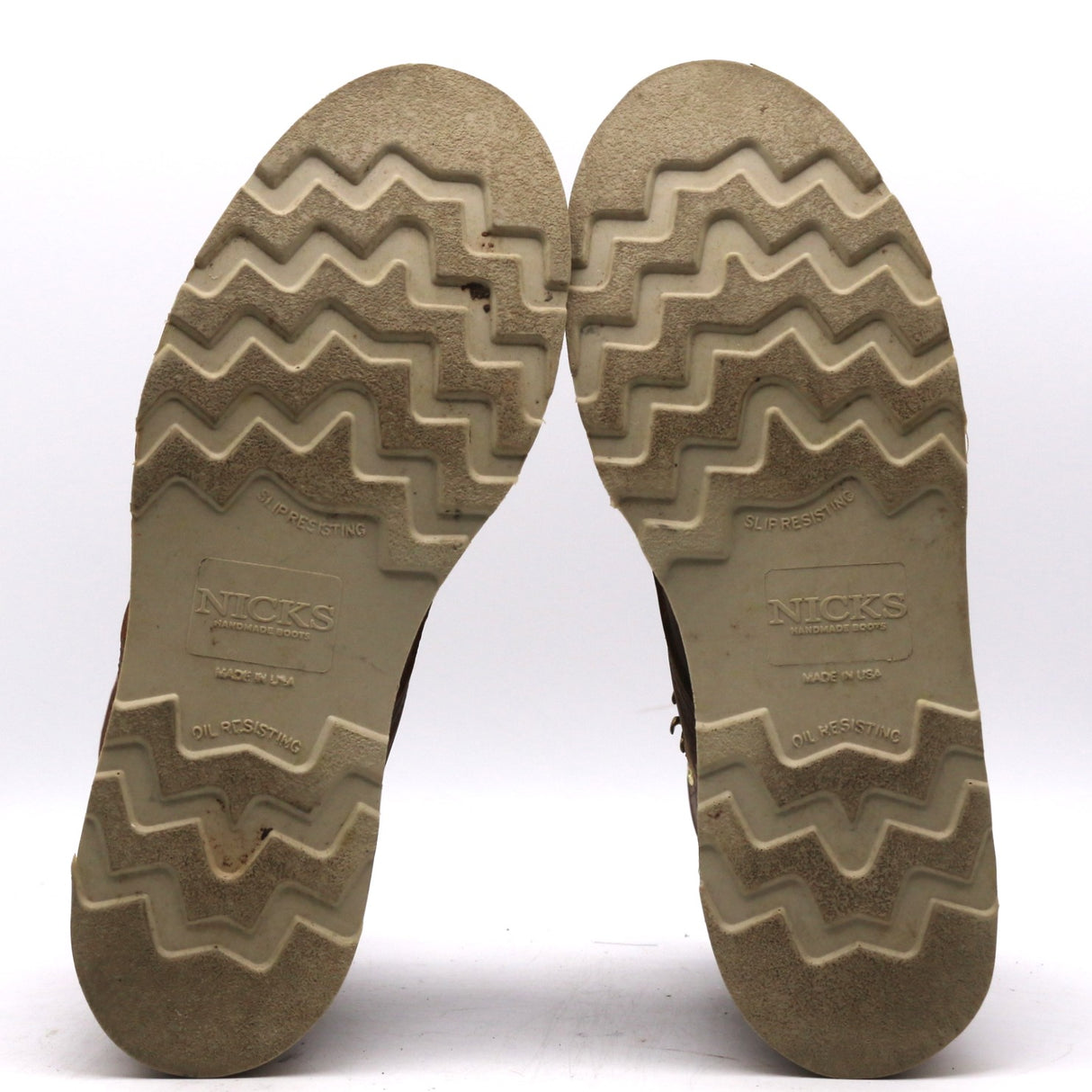 A pair of boots shown from the bottom, displaying their textured zig-zag treaded soles. The soles are a weathered tan-brown color with the brand name 'NICKS' clearly embossed in the center.