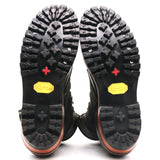 A pair of black boots shown from the sole view, featuring deep treaded rubber soles with a red cross symbol and a yellow Vibram logo, set against a plain white background.