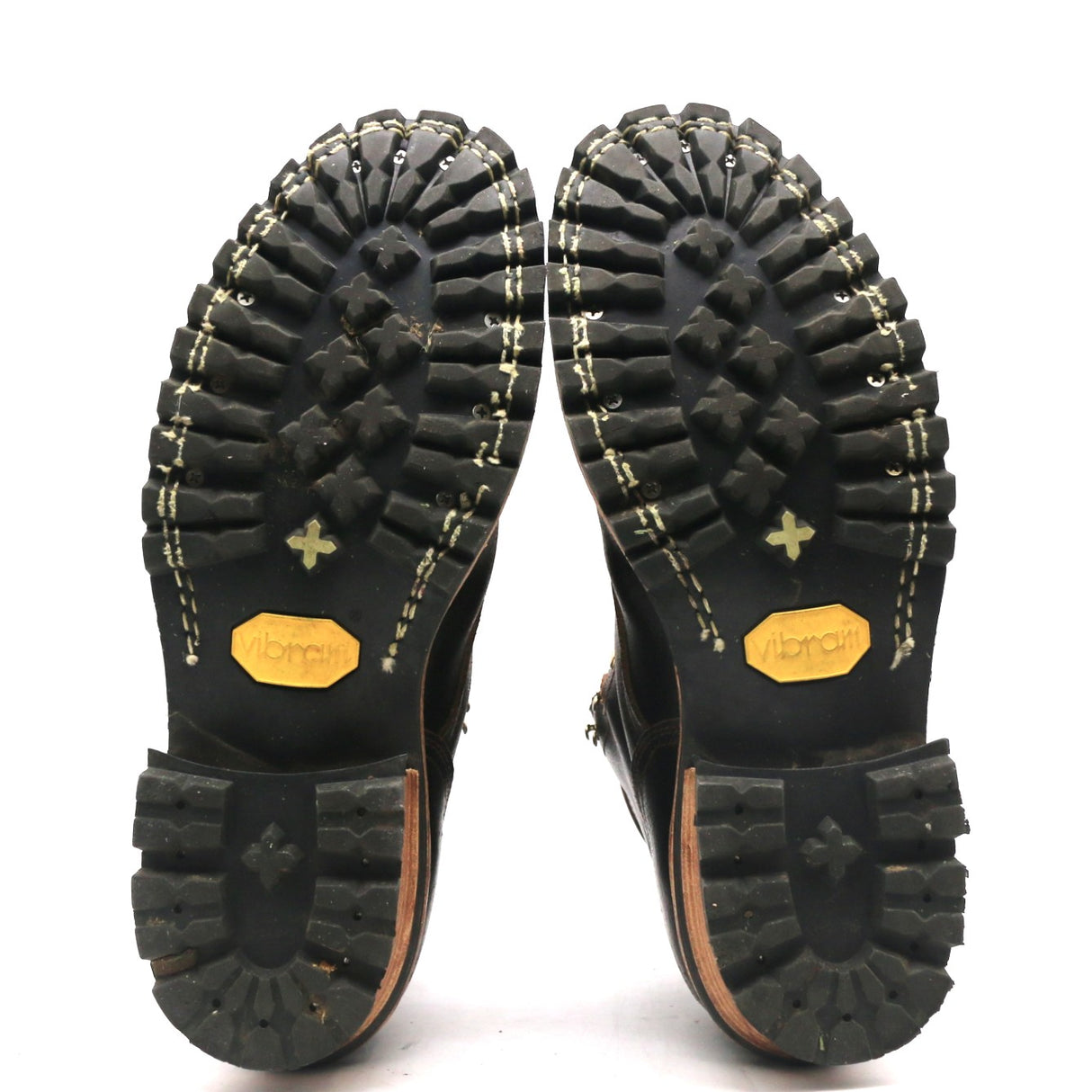 A pair of black boots shown from the sole side, displaying deep lugged rubber tread, contrast stitching, and yellow Vibram logos on the soles against a white background.