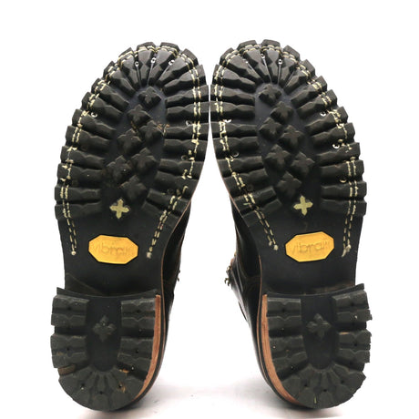 A pair of black boots shown from the sole side, displaying deep lugged rubber tread, contrast stitching, and yellow Vibram logos on the soles against a white background.