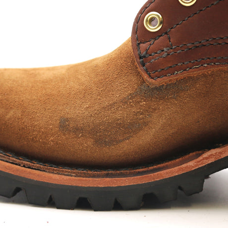 Close-up of a brown suede work boot showing the toe, side stitching, brass eyelets, and a black lug sole on a white background.