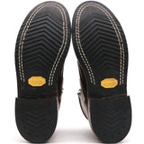 The soles of a pair of dark brown leather boots, showing the black treaded rubber outsoles with white contrast stitching and yellow Vibram logos.