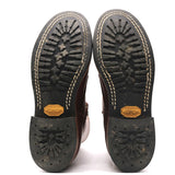 A pair of boots shown from the bottom, displaying the black rubber soles with deep tread patterns, beige contrast stitching, and gold-colored Vibram logos on the heel.