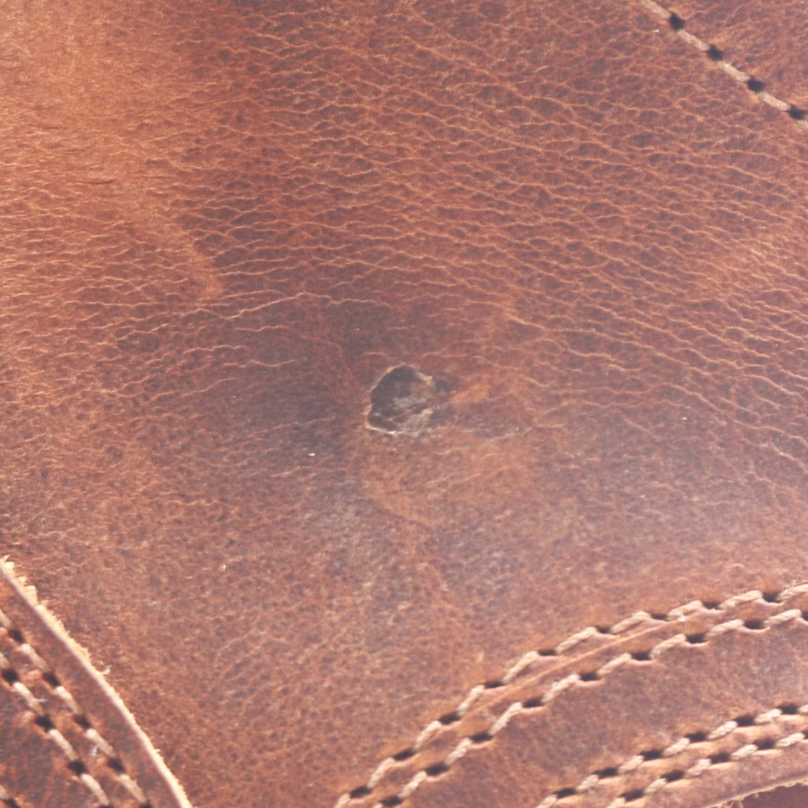 Close-up of a brown leather surface with visible grain texture, stitching, and a small dark stain or defect near the center.