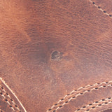 Close-up of a brown leather surface with visible grain texture, stitching, and a small dark stain or defect near the center.