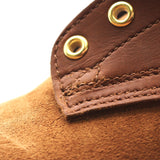 Close-up detail of a brown leather strap with brass grommets and a textured brown suede or suede-like surface below, photographed on a white background.