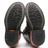 The soles of a pair of black leather boots, showing the treaded black rubber outsoles with white stitching, a black heel cap with a starburst pattern, and a tan leather midsole.