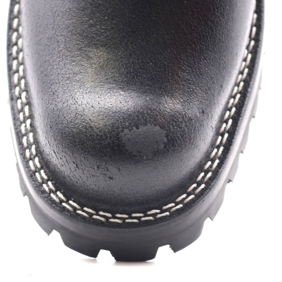 Close-up of a black leather boot toe showing white contrast stitching and a black treaded sole on a white background.