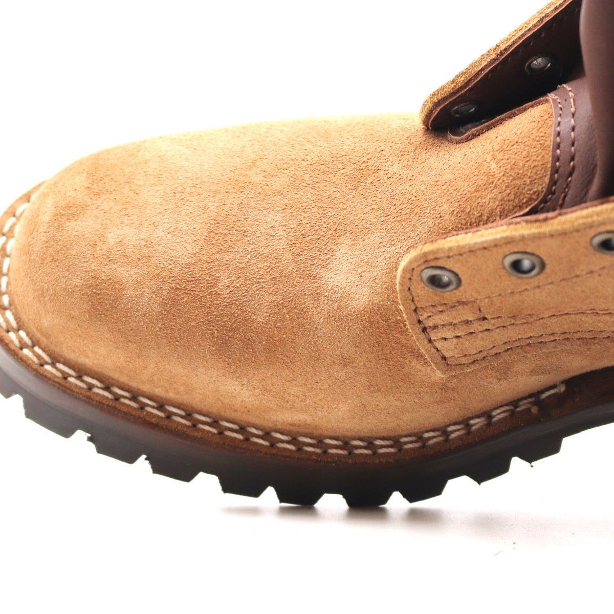 Close-up of a tan suede work boot with dark brown laces, metal eyelets, and a black lug sole on a white background.