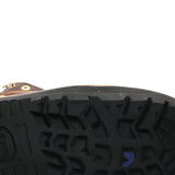 Close-up of the treaded black rubber sole of a brown boot, showing a small blue chevron logo and a gold grommet on a white background.