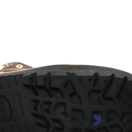 Close-up of the treaded black rubber sole of a brown boot, showing a small blue chevron logo and a gold grommet on a white background.