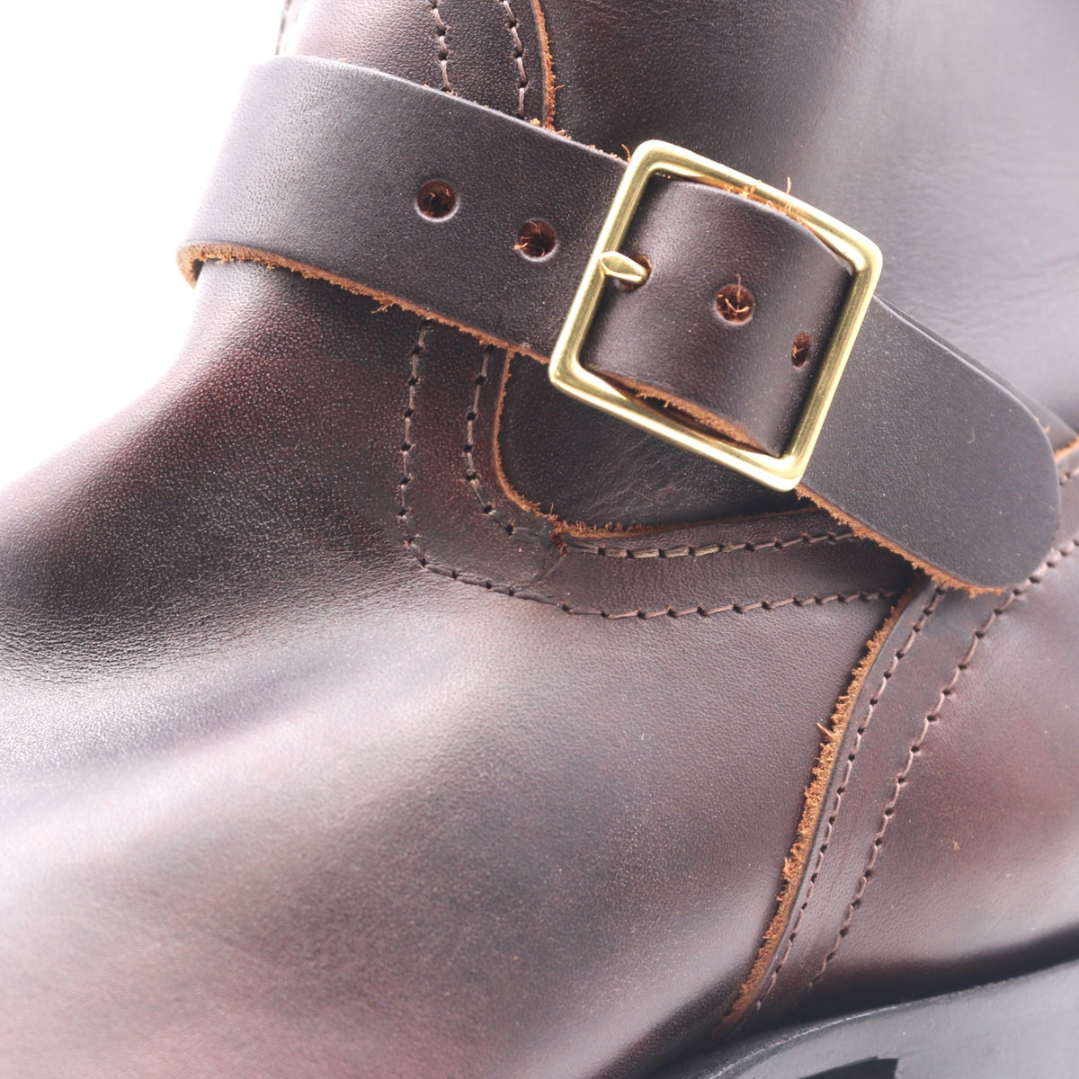 Close-up of a dark brown leather boot strap with a gold-tone metal buckle, showing detailed stitching and texture.