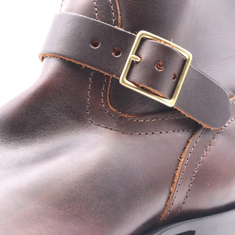 Close-up of a dark brown leather boot strap with a gold-tone metal buckle, showing detailed stitching and texture.