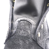 Close-up detail of a black leather and suede boot showing the stitching and a gold-tone metal rivet on a white background.