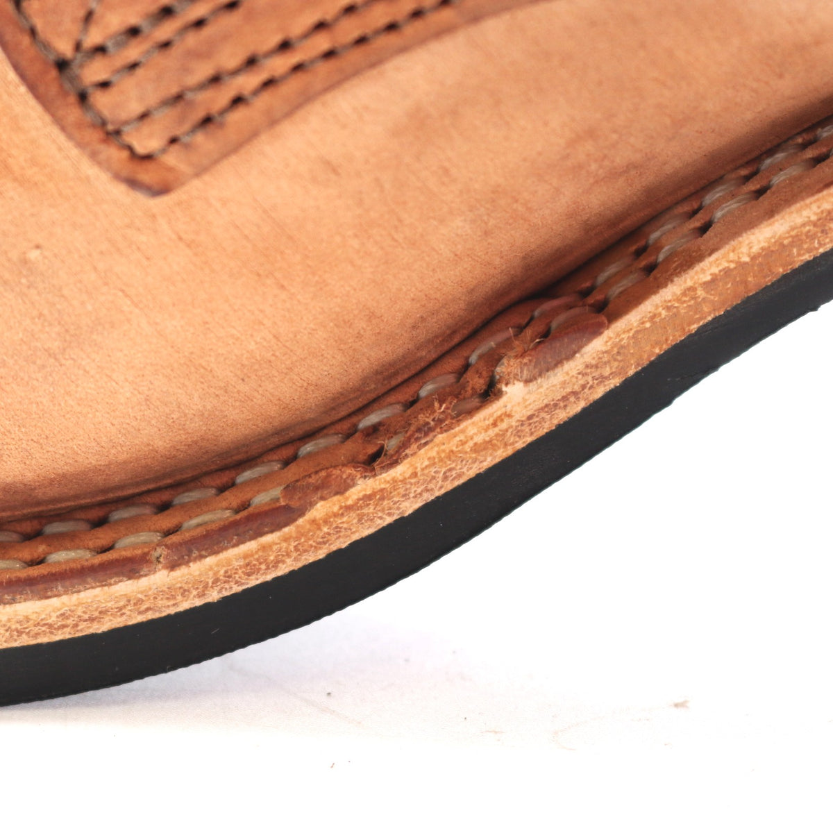 Close-up of a light brown leather shoe corner showing the stitched vamp, layered leather edge, and black outsole against a white background.