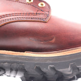 Close-up of a brown leather boot showing the side panel, stitching, brass eyelets, and a black rugged sole.