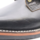 Close-up of a black leather boot toe showing scuffed leather texture, white stitching, a brown midsole layer, and part of a brown pull tab with a brass grommet.
