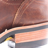 Close-up of a brown leather boot showing the side panel, stitching, and a worn stacked rubber sole with exposed layers.