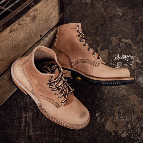 A pair of light tan suede lace-up boots with brown laces and black soles, styled on a dark rustic wooden floor next to a wooden crate.