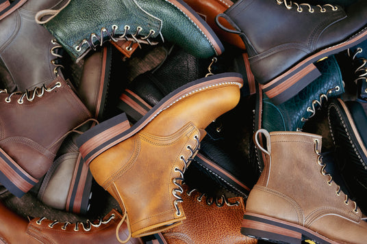 Pile of handcrafted mens lace-up leather boots in various colors including tan brown black and green